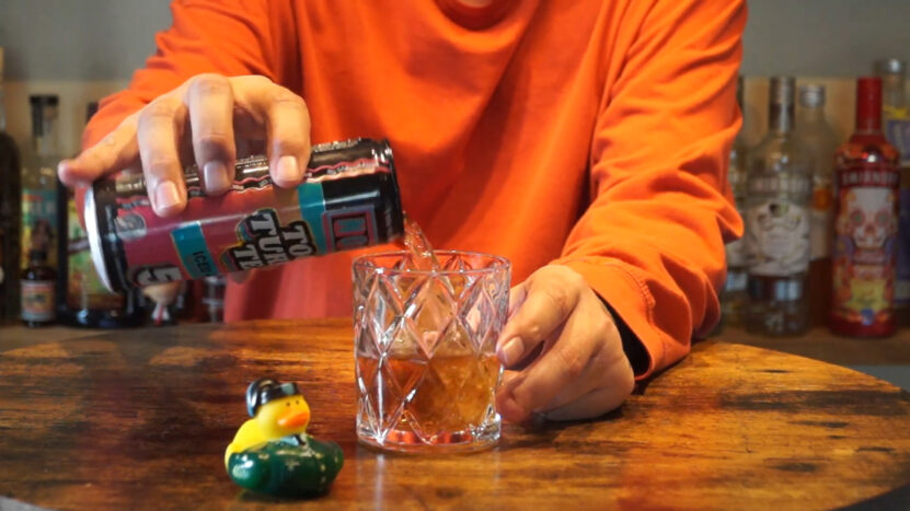 person in an orange shirt pouring a drink from a can labeled TooTurnt Tea into a glass, with a rubber duck in the foreground